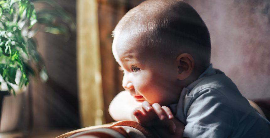 Little, cheerful two years old boy looking out the window and thinking and waiting for something. Sun shine